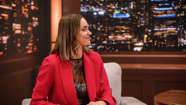 Late Night Talk Show Interview: Female Celebrity Guest Sits in Armchair Smiling Warmly at Host Presenter Off Camera During a Fun Chat, Glowing Night City Backdrop on Television Studio Set