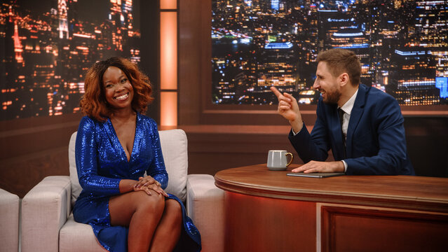 Late Night Talk Show Interview: White Male Host Points Playfully at the Black African American Female Celebrity Guest Laughing in the Armchair, Joking Together on the City Night Set