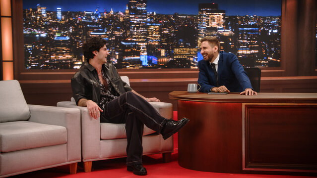 Late Night Talk Show Interview: Male Celebrity Guest Shares a Funny Story on the Couch While the Smiling Host Laughs Behind His Desk, Sparkling Night City Skyline Lighting the Studio Set