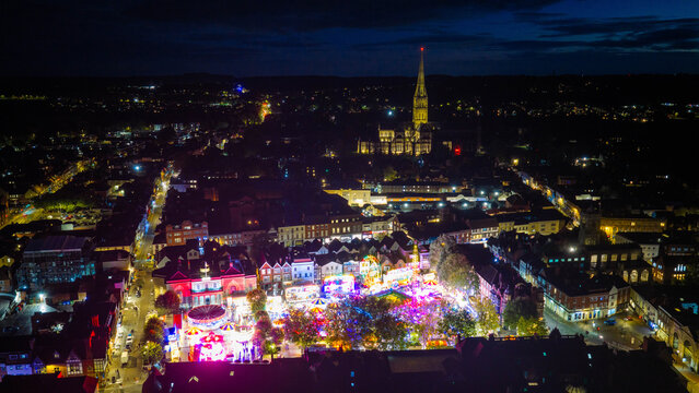 Aerial view of Salisbury Charter Fair illuminated at night with colorful carnival rides, set against the historic city skyline and Salisbury Cathedral, Salisbury, England, United Kingdom.