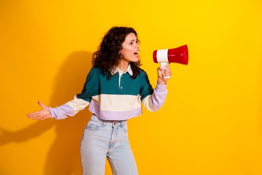 Young woman shouting into a red megaphone against a bright yellow background expressing urgency, protest and strong emotion in casual jeans and a color-block sweatshirt
