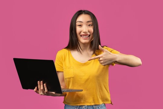 Smiling Young Asian Woman Pointing At Laptop While Holding It On Pink Background, Showing Excitement And Happiness. Setting Is Studio With Business Concept And Digital Technology Emphasis.