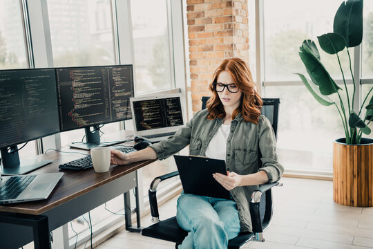 Redhead software developer reviewing code on a clipboard in a bright modern home office with multiple monitors and laptop while holding a coffee mug