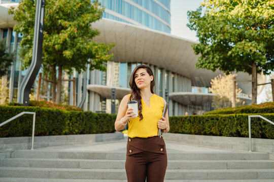 Confident businesswoman holding coffee and laptop in city