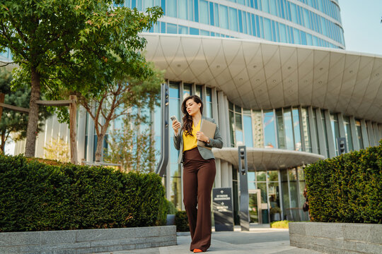 Professional businesswoman walking while using smartphone outside office building