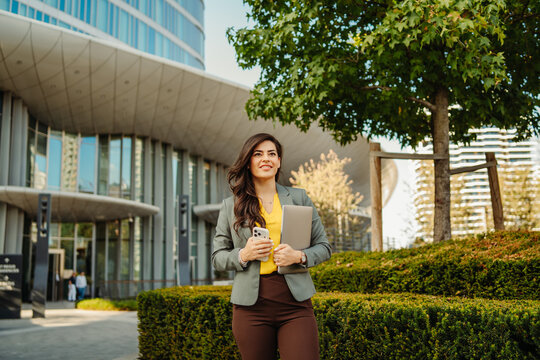 Confident businesswoman walking with laptop and phone outdoors