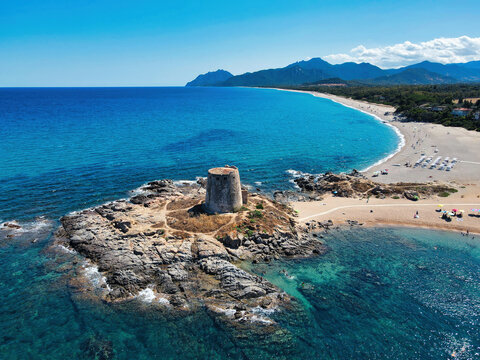 Aerial view of the ancient watchtower of Bari Sardo on a rocky headland, with blue sea, sandy beach and mountains in the background, Sardinia, Italy
