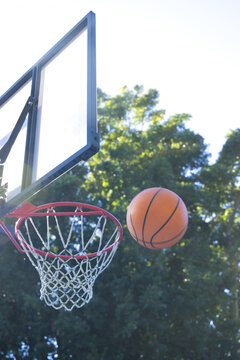 Orange basketball is arcing toward red-rim hoop with clear backboard near leafy park trees