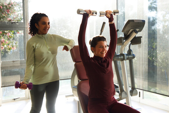 Diverse female trainer-client lifting and spotting on bench in gym with dumbbells in sportswear
