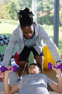 African American coach spotting Asian female client lifting purple dumbbells on bench by windows