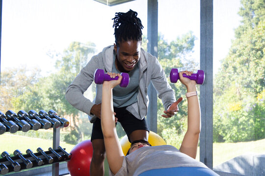 Male trainer in workout clothes spotting female client pressing purple dumbbells on bench in studio