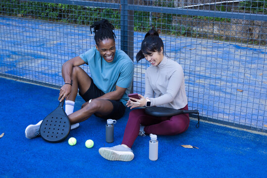 Diverse friends sitting on bright blue racket court near fence, sharing phone, paddles, neon balls