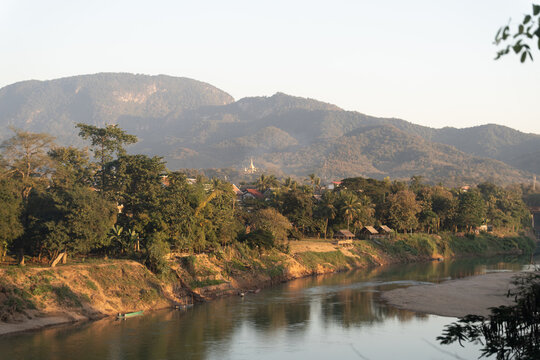 Aerial view of Nam Khan River flowing through lush tropical vegetation with small wooden huts along the riverbank and mountains in the background, Luang Prabang Province, Laos.