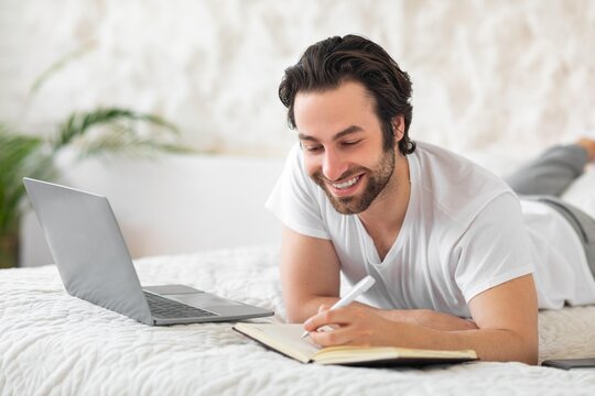 Smiling Young Caucasian Man Enjoying Time While Writing In Notebook, Lying On Bed With Laptop Open In Cozy Home Setting, Reflecting On Ideas And Plans For Future Projects, Business Concept.