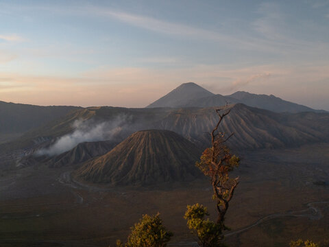 Aerial view of Mount Bromo with its volcanic crater emitting smoke, surrounded by the vast sandy caldera and rugged mountain landscape under a soft morning sky in Tosari, East Java, Indonesia.