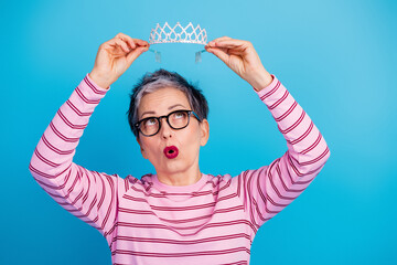 Playful mature woman holding a tiara above her head expressing surprise and confidence against a bright blue background © deagreez