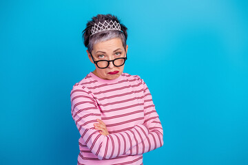 Unimpressed middle-aged woman wearing a tiara and glasses with crossed arms against a bright blue background © deagreez