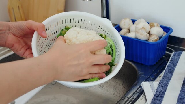 Person washes fresh cauliflower head under tap water using white colander in kitchen sink, thoroughly rinsing healthy raw vegetable for nutritious meal preparation with mindful culinary care.