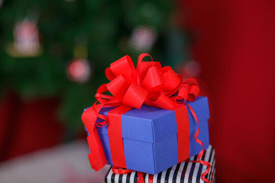 Blue gift box with large red ribbon bow stacked on striped present against blurred Christmas tree and red background, holiday celebration and giving concept