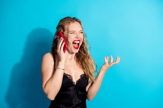 Excited young woman talking on a red smartphone, laughing and shouting against a bright blue background in a black silk camisole &mdash; expressive lifestyle portrait of communication and joy
