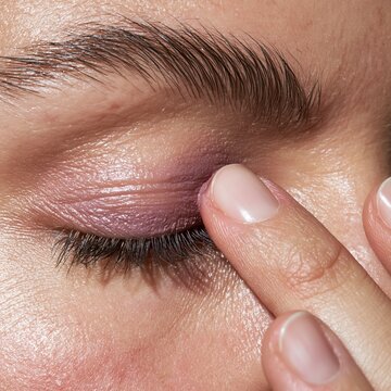 a young woman gently applying soft mauve cream eyeshadow onto her upper eyelid with her index finger pad