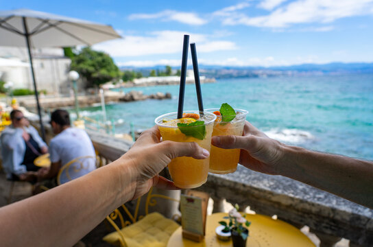 Two hands clink summer cocktails by the sea at a sunny outdoor cafe