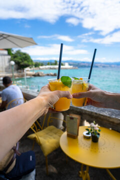 Two hands clink summer cocktails by the sea at a sunny outdoor cafe