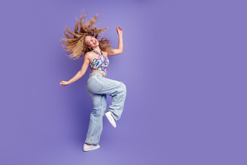 Naklejka na ściany i meble Happy young woman jumping and dancing in casual jeans and colorful crop top on purple background, carefree energetic lifestyle portrait expressing joy and movement