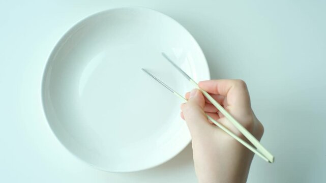 A female hand holds metal chopsticks against the backdrop of empty white plate, top view.