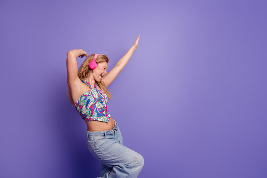 Happy young woman dancing with pink headphones against a purple background, enjoying music and carefree summer style in colorful crop top and relaxed denim jeans