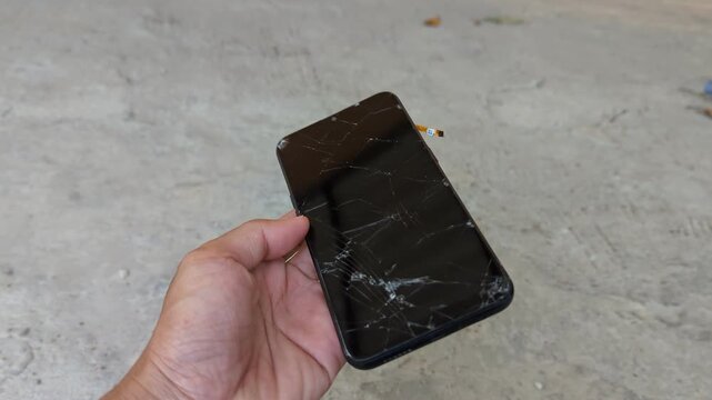 A person's hand holding a damaged smartphone with a heavily cracked glass screen against a blurry concrete background.