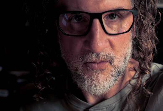 Dramatic close-up portrait of a mature man with curly hair, gray beard and black glasses, half of his face in shadow, moody low key indoor lighting