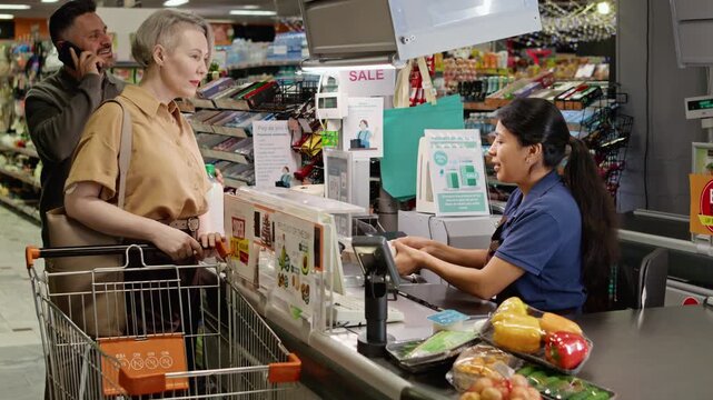Smiling Hispanic cashier scanning groceries for middle aged Caucasian woman at supermarket checkout counter. Ideal for retail service, grocery store and consumer banking commercials