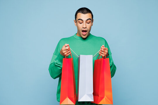Young shocked surprised man he wearing green casual clothes hold in hand open look at paper package bag after shopping isolated on plain pastel blue cyan background. Black Friday sale buy day concept