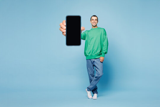 Full body young middle eastern man wear green shirt casual clothes hold in hand use mobile cell phone with blank screen workspace area isolated on plain light blue cyan background. Lifestyle concept.