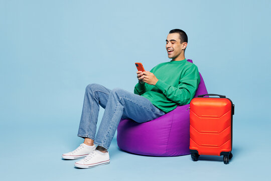 Traveler happy man wear casual clothes sit in bag chair near suitcase bag use mobile cell phone isolated on plain blue background. Tourist travel abroad in free time getaway. Air flight trip concept.