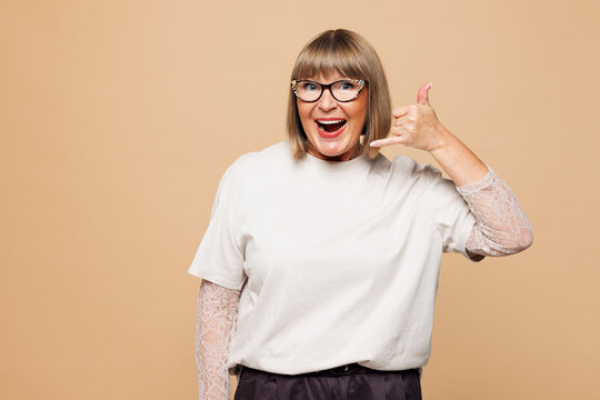 Elderly blonde woman 50s years old she wears white shirt casual clothes doing phone gesture like says call me back isolated on plain pastel light beige background studio portrait. Lifestyle concept.