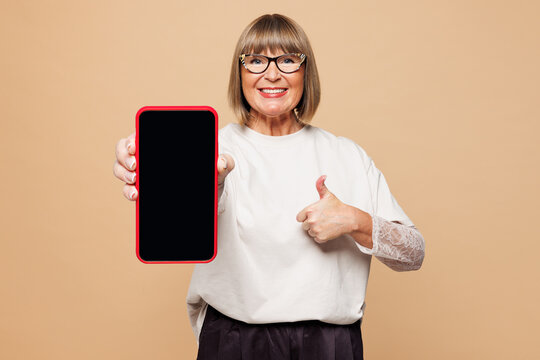 Elderly blonde woman 50s years old she wear white shirt casual clothes hold use blank screen mobile cell phone show thumb up isolated on plain pastel light beige background studio. Lifestyle concept.
