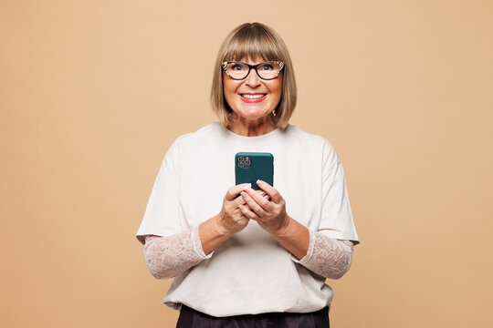 Elderly smiling happy blonde woman 50s years old she wearing white shirt casual clothes holding in hand use mobile cell phone isolated on plain pastel light beige background studio. Lifestyle concept.
