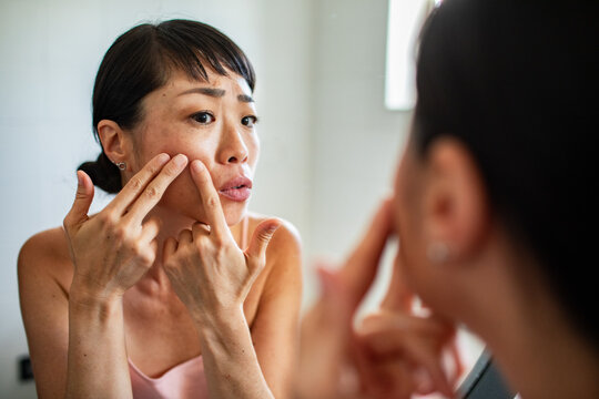 Woman squeezing pimple on cheek in bathroom mirror