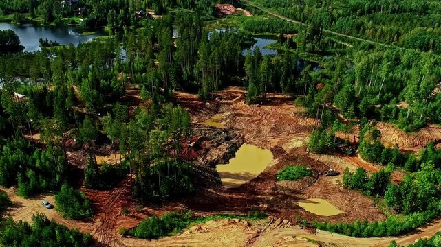 Aerial view of a forest clearing featuring active excavation work, muddy terrain, and small pools of water surrounded by dense pine trees in Latvia.