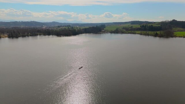 Eindrucksvolles Drohnenvideo ueber Waginger See in Suedostbayern. Einzelner Fischer in kleinen Boot ist auf weiten See zu sehen, umgeben von bewaldeten Huegeln und einer friedlichen Naturlandschaft.