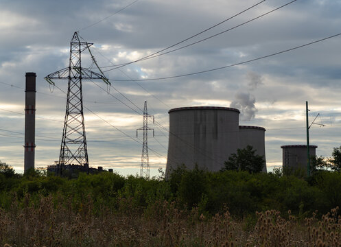 View of cooling towers, a smokestack and transmission lines at an industrial power station in Visonta, Hungary, beneath a cloudy summer sky and surrounded by wild vegetation.