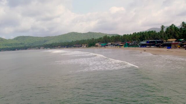 Aerial view of the sandy shoreline and rolling waves at Vagator Beach with lush green hills and palm trees in the background in Anjuna, Goa, India.