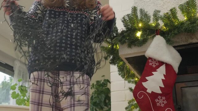 Person in a cozy sweater and plaid pajama pants untangles Christmas string lights near a fireplace with a stocking. Cozy holiday preparation at home