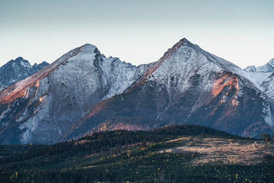 View of Belianske Tatras mountains with snow-capped peaks, rugged rocky slopes, and dense pine forests under a clear sky during golden hour in Lapszanka, Lesser Poland Voivode, Poland.