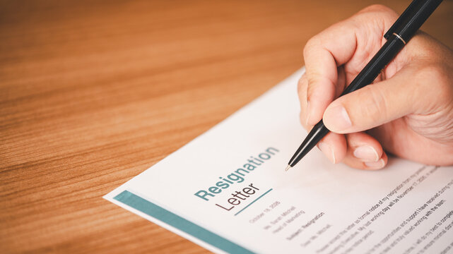 Person hand writing a resignation letter on a wooden desk with a black pen. Career change, job quitting, employment termination, and corporate office transition concept
