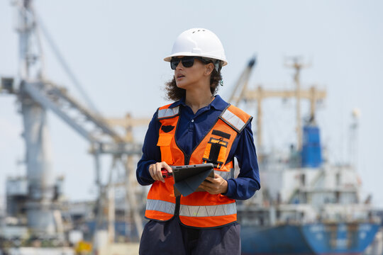 Confident female engineer holding tablet inspects industrial commercial port terminal wearing safety vest hardhat and sunglasses