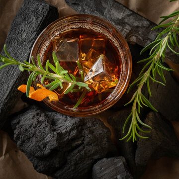 Old fashioned cocktail in a whisky glass with a sprig of fresh rosemary and orange peel, sitting on a bed of chunk charcoal, from above