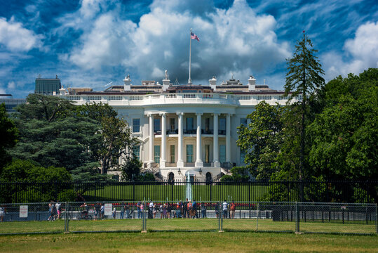 View of the White House with its neoclassical architecture and white facade, set against a blue sky, surrounded by trees and a fence in Washington, District of Columbia, United States.